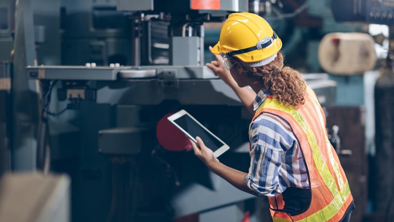 A female factory worker wearing hard hat and safety glasses inspecting a machine on the plant floor while holding a tablet