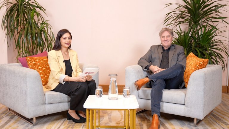 Two people in business attire seated in white armchairs, with a table and two cup of water.