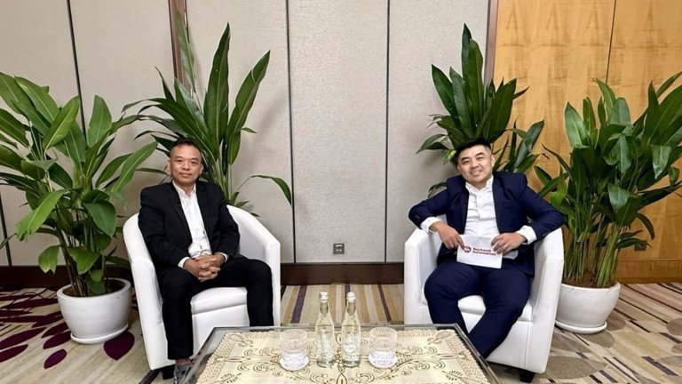 Two people in business attire seated in white armchairs, with a table and large potted plants in an indoor setting.