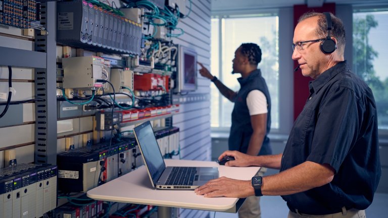 Two male technical support engineers in Rockwell Automation remote support center mount FLEXHA 5000 i/o modules while talking to customer on headset.Rename file as: two-male-remote-support-engineers-work-on-FLEXHA-5000-modules.RA-6293