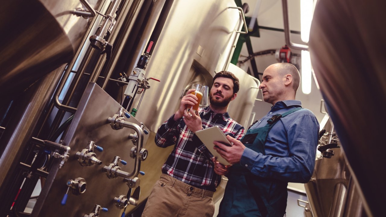 Two man standing in brewery with one holding a glass of beer