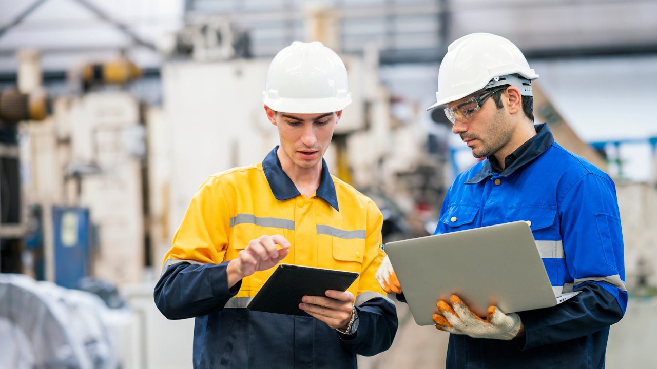 Two people in hardhats are looking at their tablet and laptop and having a discussion in a factory