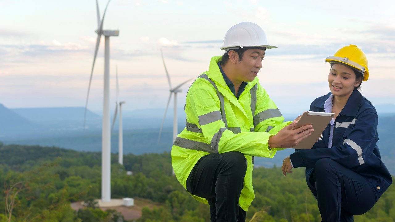 Two people in hardhats looking at a digital tablet near wind turbines