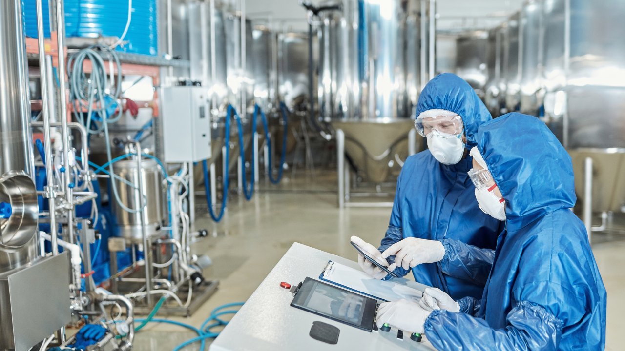 Two people in PPE examining data from chemical processes in stainless steel vats in the room. Side view portrait of two workers operating equipment at factory and using control panel
