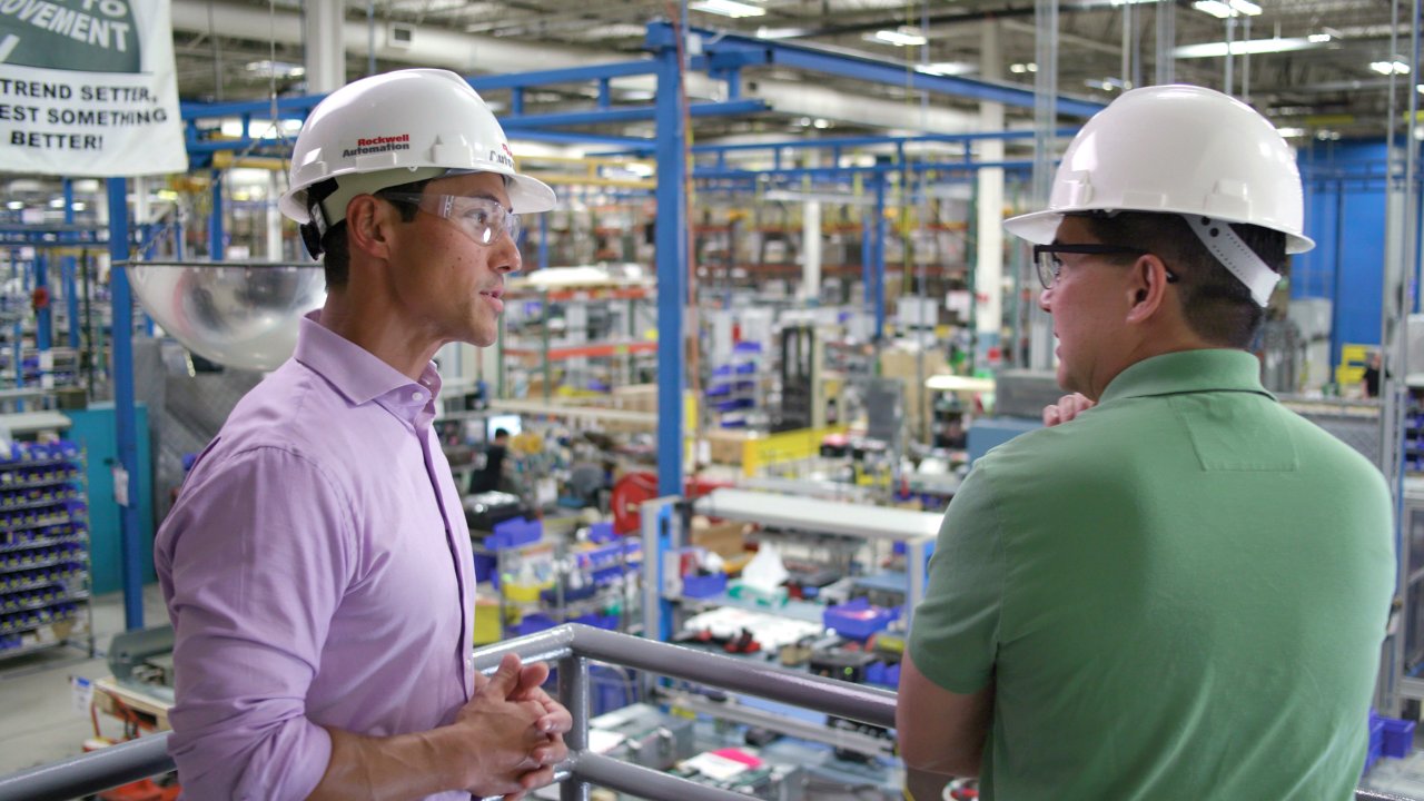 A Rockwell Automation industrial asset management specialist in white hard hat and button-down business shirt advises a customer wearing a green polo shirt on maintenance, repair and asset optimization strategies as they overlook factory floor. 