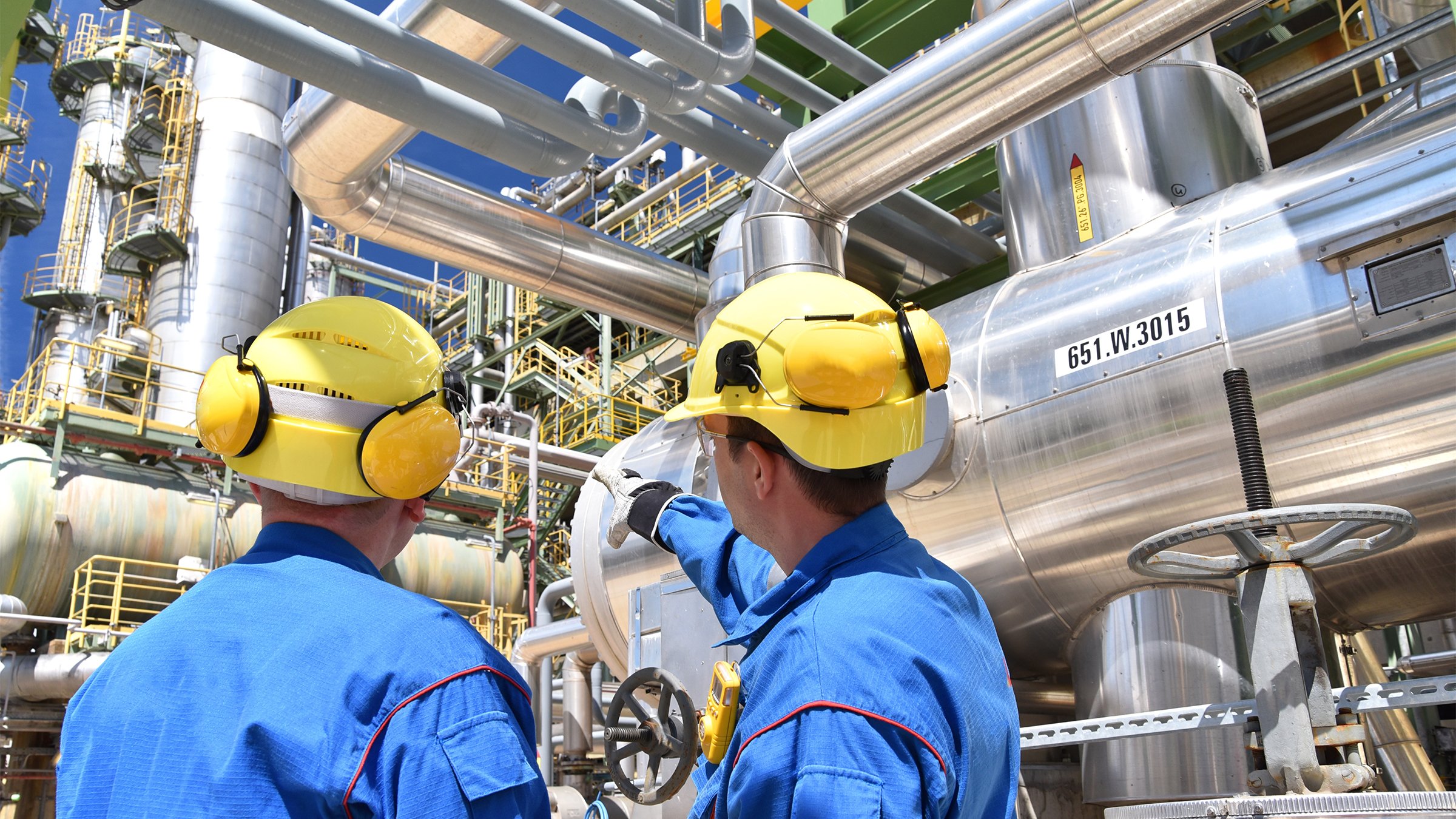 Two industrial workers in blue uniforms outside an oil processing refinery looking at pipes and equipment.