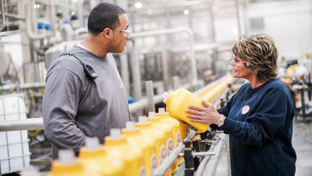 Due dipendenti sulla linea di produzione di Church & Dwight. Un uomo e una donna, indossano entrambi occhiali protettivi. Dipendente donna che preleva una bottiglia gialla di detergente dal nastro trasportatore.