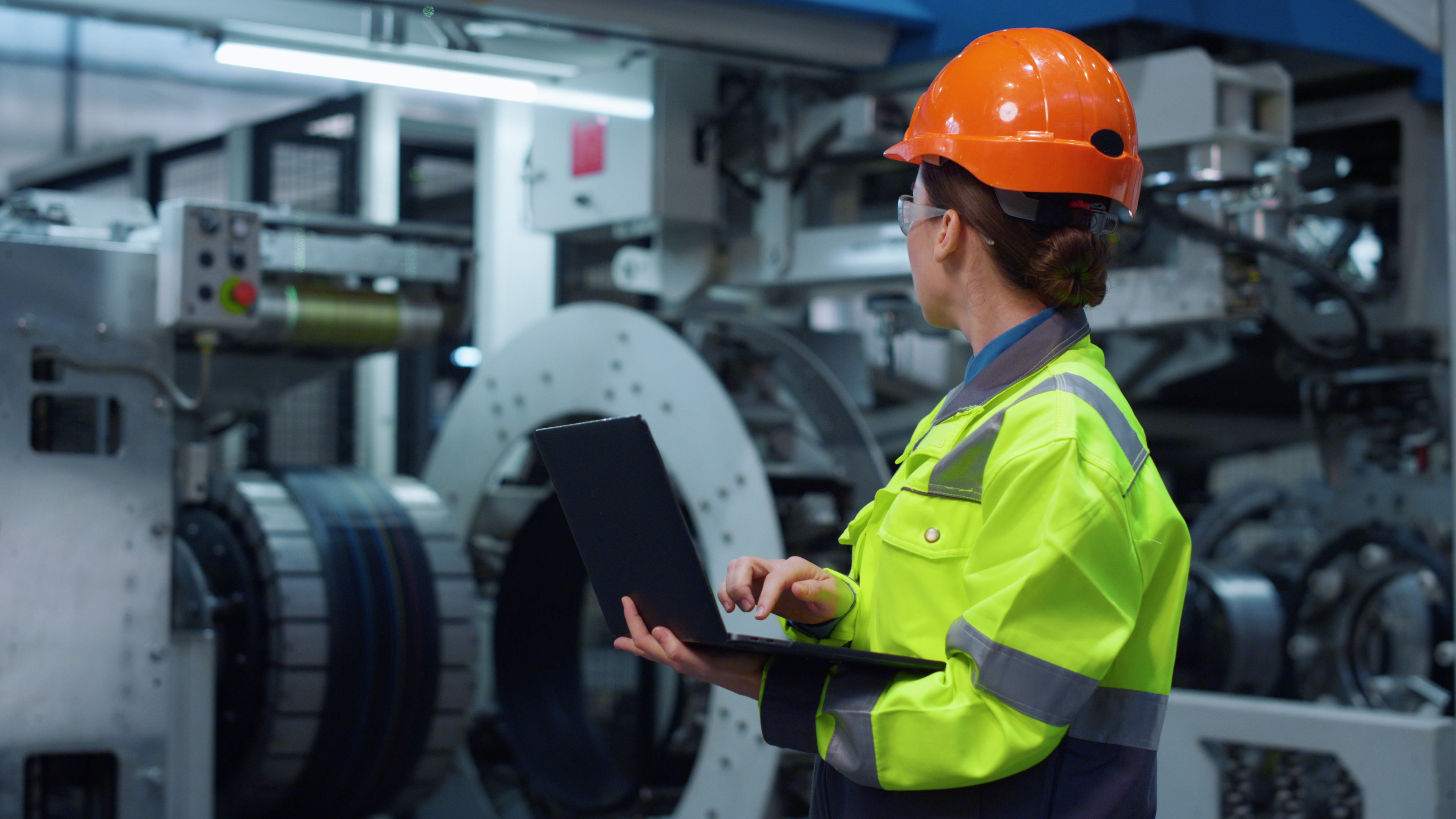 Brunette specialist working laptop computer on modern huge digital factory. Industrial specialist typing laptop computer wearing safety hardhat in manufacturing facility. Professional concept.