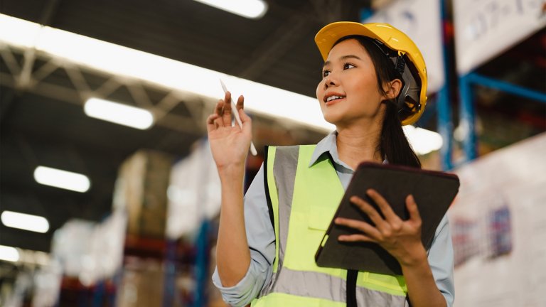 Female wearing yellow hardhat, holding clipboard in warehouse, managing OTTO autonomous mobile robot (AMR robot) fleet