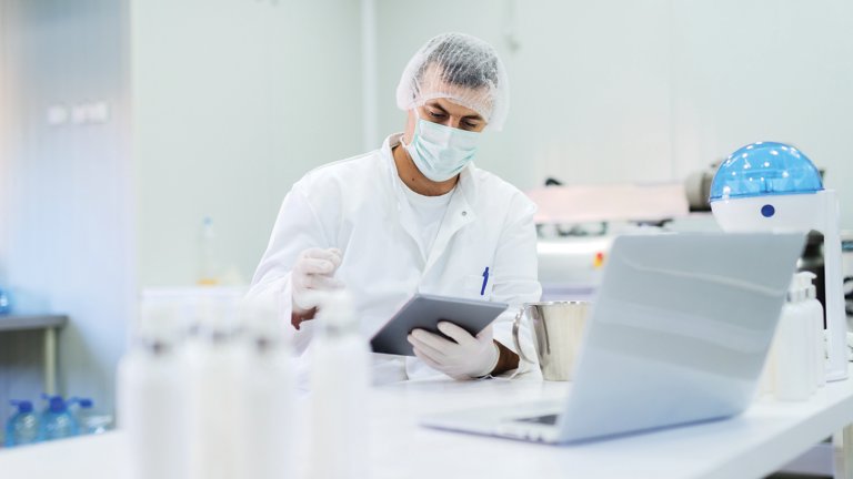 Imagem de um homem vestindo roupas esterilizadas sentado em um laboratório iluminado e verificando a qualidade dos produtos, enquanto segura o tablet em suas mãos e lê anotações.