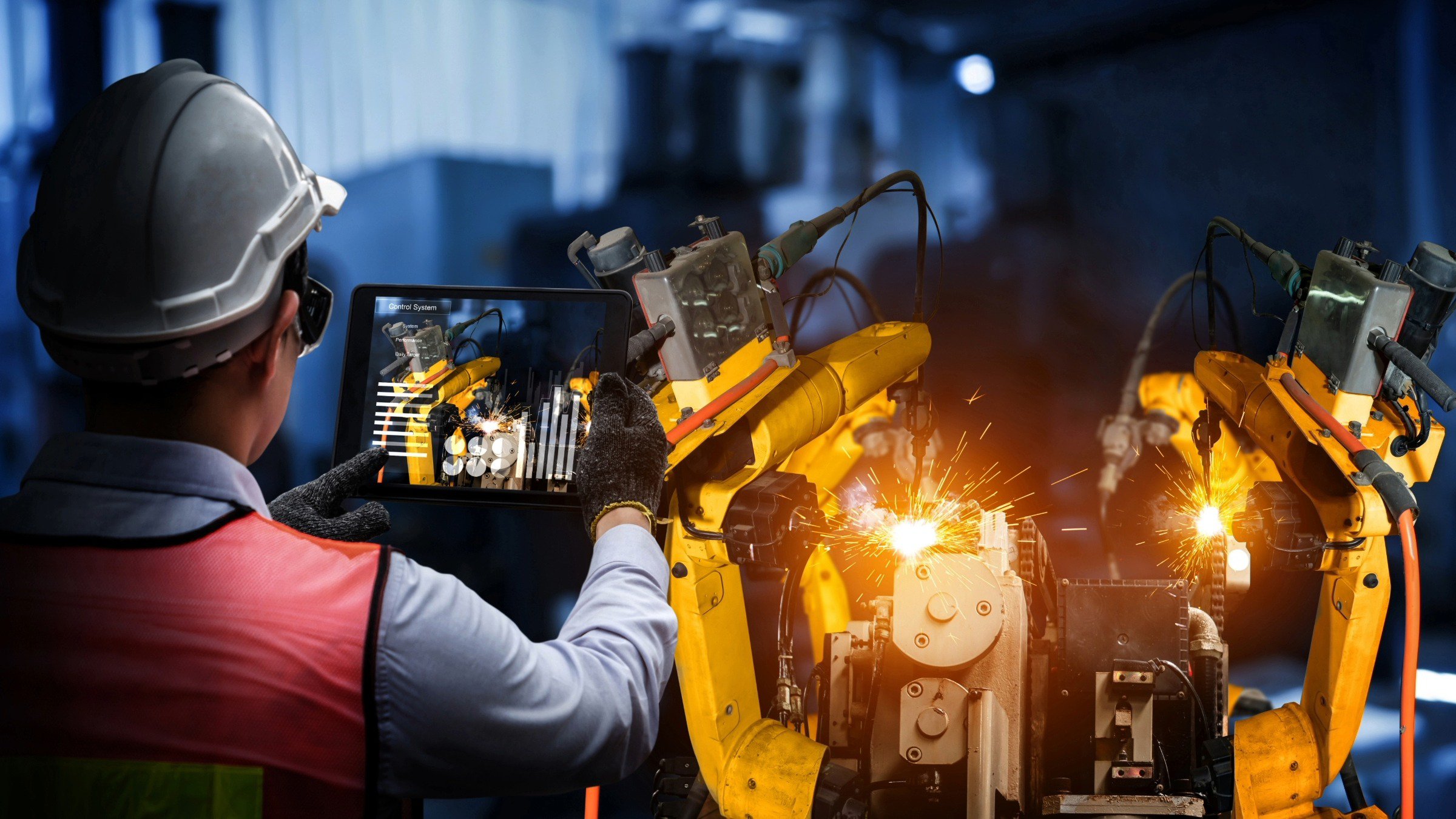 Worker with safety vest using a tablet to focus on robotic manufacturing process.