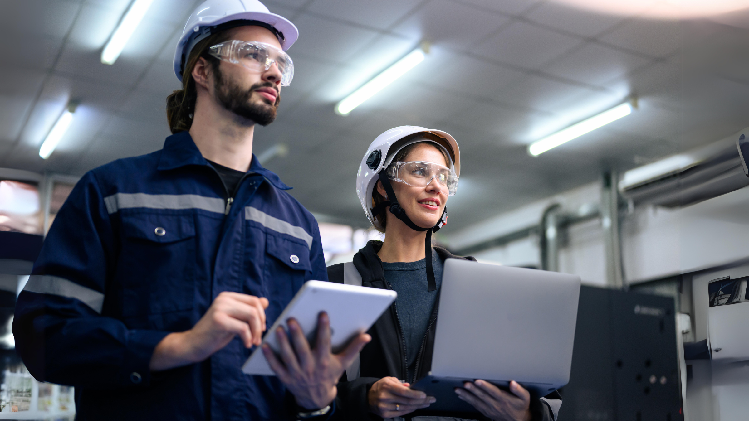 Dos trabajadores con cascos y gafas de seguridad, sosteniendo computadoras portátiles mientras trabajan juntas