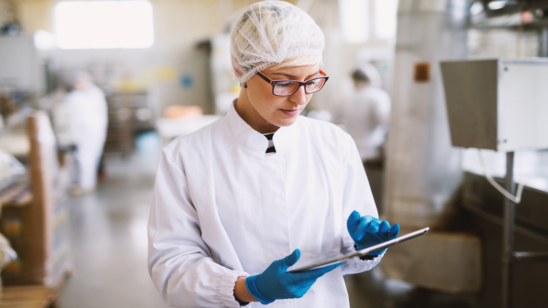 Employee in food manufacturing plant wearing hairnet and gloves and holding digital tablet.