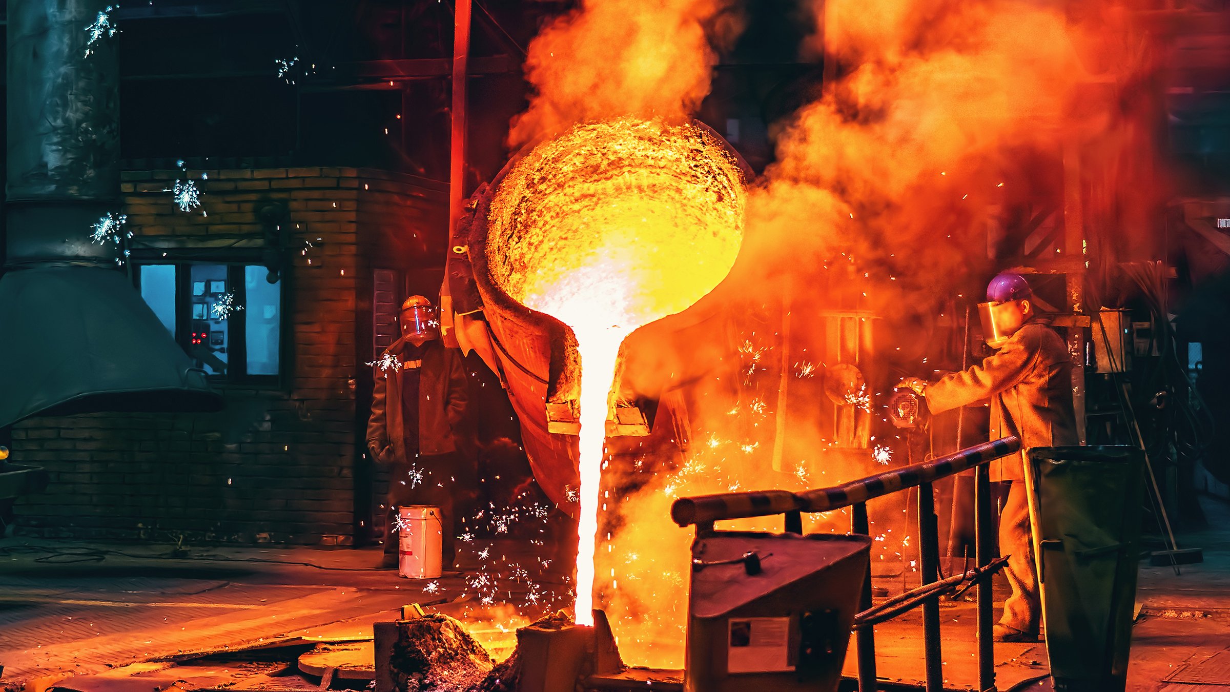 Bright white and yellow hot molten steel is poured out of a large bucket in a steel foundry. White sparks and orange smoke surround the activity.