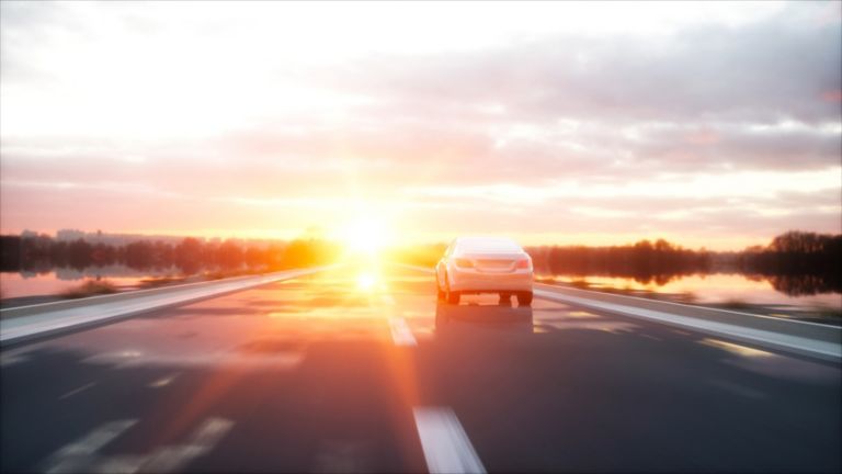 A rear view of a car on a roadway driving toward the sun on the horizon