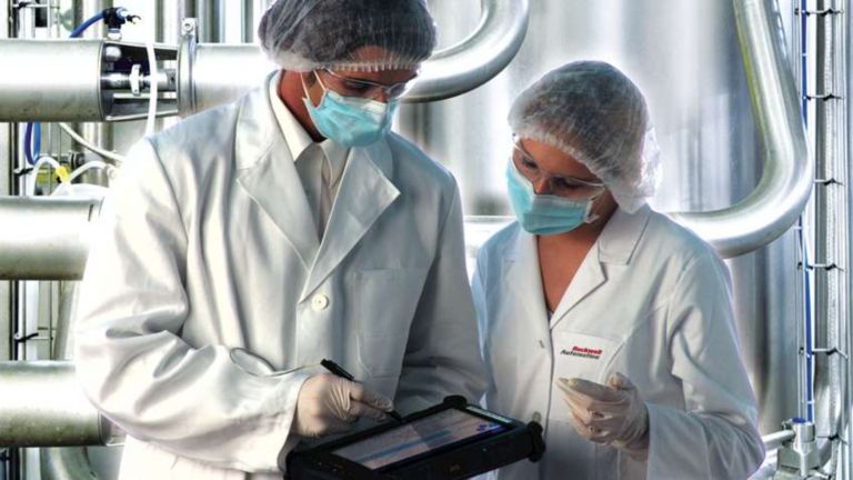 머리망, 마스크, 흰 장갑, 실험실복을 입은 두 명의 직원이 공장에서 태블릿으로 정보를 보고 있습니다.