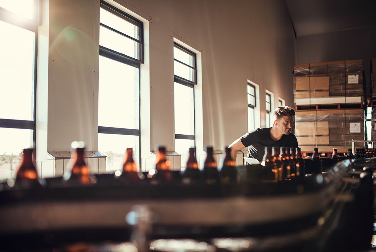 Young man supervising the production of craft beer at brewery. Man working at alcohol manufacturing factory.