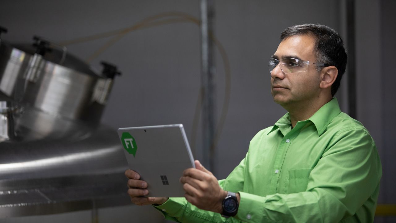 Male employee wearing a green shirt and safety glasses holding a tablet with a green FactoryTalk logo assessing process data