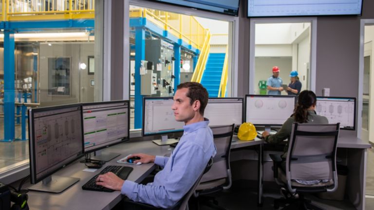 Ein Mitarbeiter sieht sich die Messdaten auf seinem Monitor an, während ein anderer Mitarbeiter die Daten in einem anderen Büro mit Fenstern zur Sicht auf die Produktionshalle auswertet