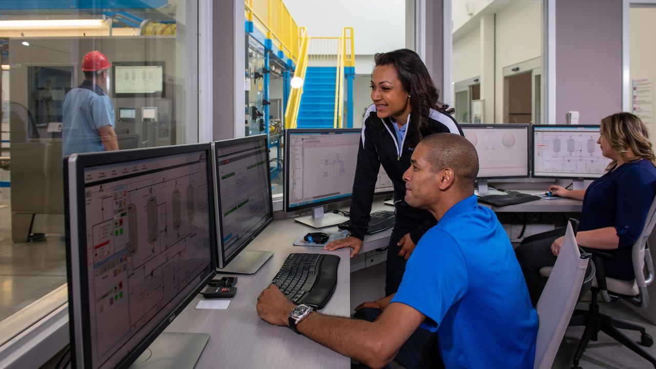 Three smiling employees viewing FactoryTalk Transaction Manager software on a monitor in a separate office with windows to view the plant floor