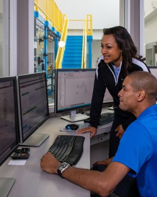 Three smiling employees viewing FactoryTalk Transaction Manager software on a monitor in a separate office with windows to view the plant floor