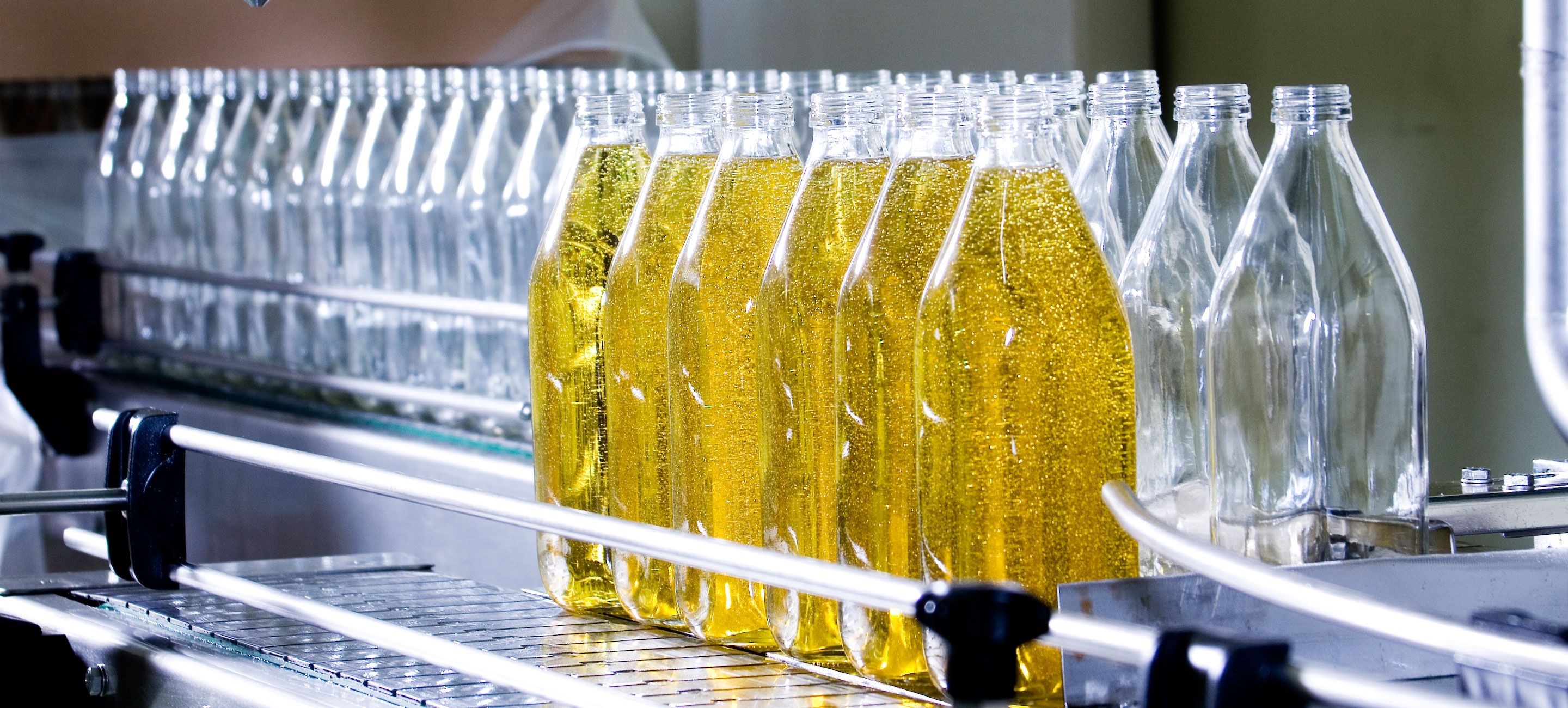 A production line of clear glass bottles, some filled with golden liquid and some empty, moving along a conveyor belt in a brightly lit factory.
