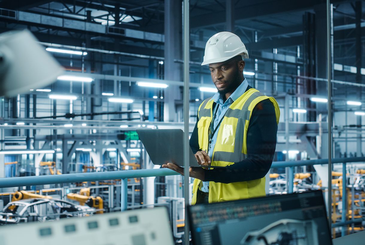 Car Factory: Professional Male Automotive Engineer Wearing Hard Hat, Walking, Using Laptop. Monitoring, Control, Equipment Production. Automated Robot Arm Assembly Line Manufacturing Electric Vehicles