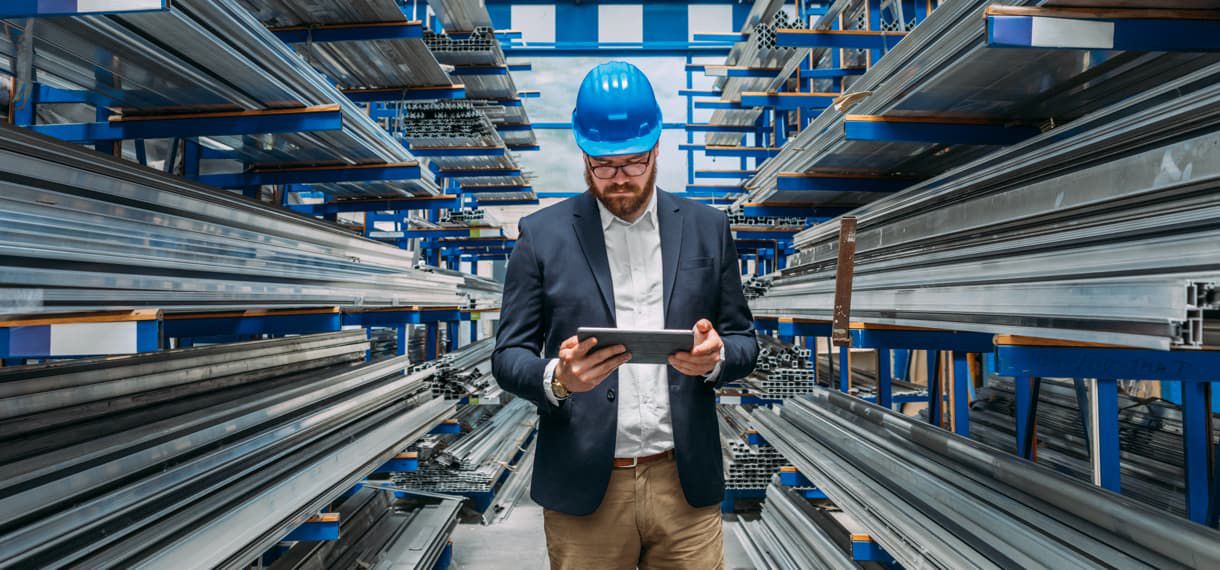 Man in blue hardhat wearing office jacket viewing digital tablet while in warehouse
