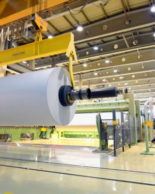 Employee looking at a paper roll on a crane for the production of paper in a modern factory
