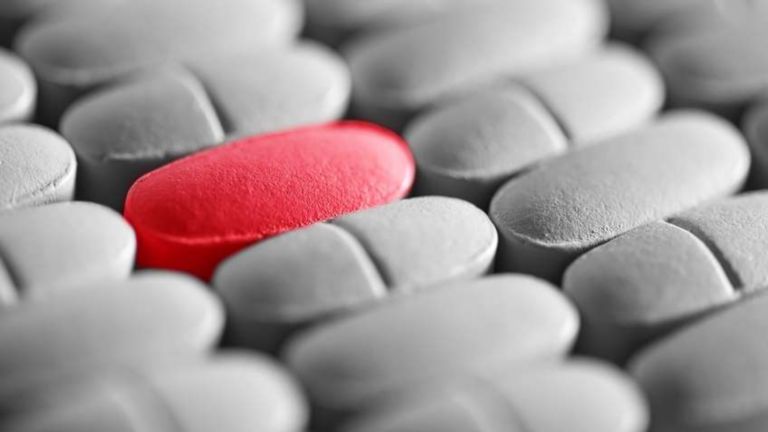 A single red pill stands out among several white pills on a pharmaceutical manufacturing line.