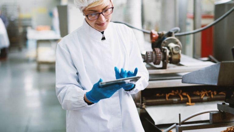 Female employee wearing a hairnet, lab coat and blue rubber gloves in a laboratory typing information into a tablet.