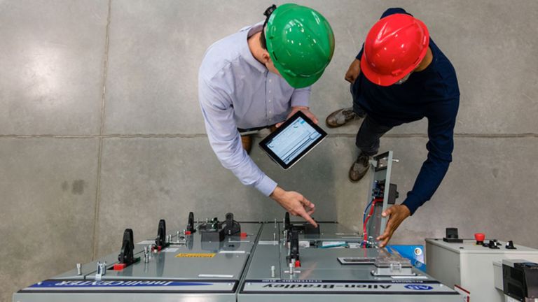 Vue aérienne de deux hommes équipés de casques regardant un appareil devant un centre de commande de moteur dans une usine.